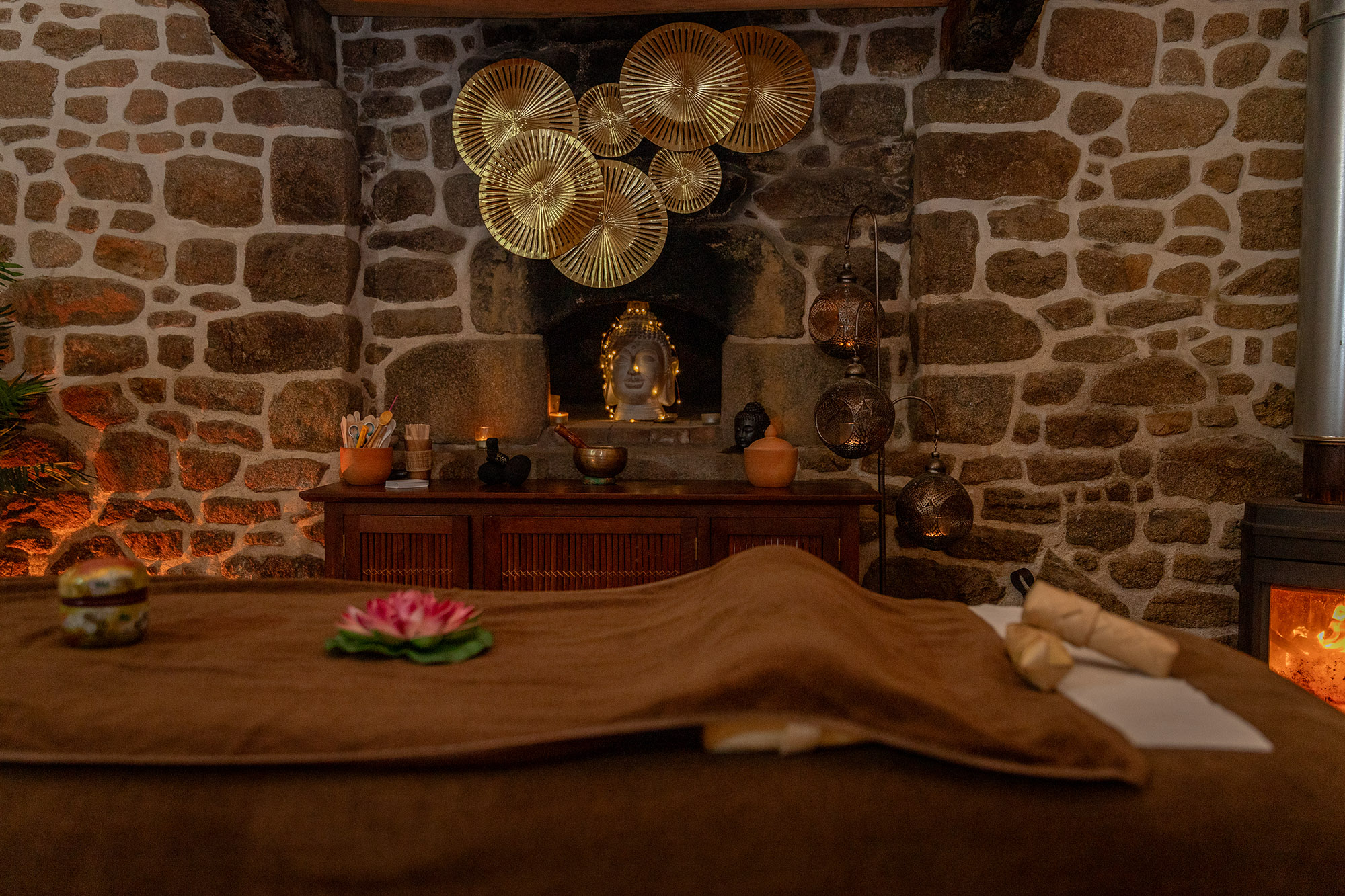 A serene spa setting featuring a massage table with a brown cloth, adorned with a pink lotus flower. The background includes a stone wall, golden decorative plates, a statue of a Buddha, and warm lighting creating a relaxing atmosphere.